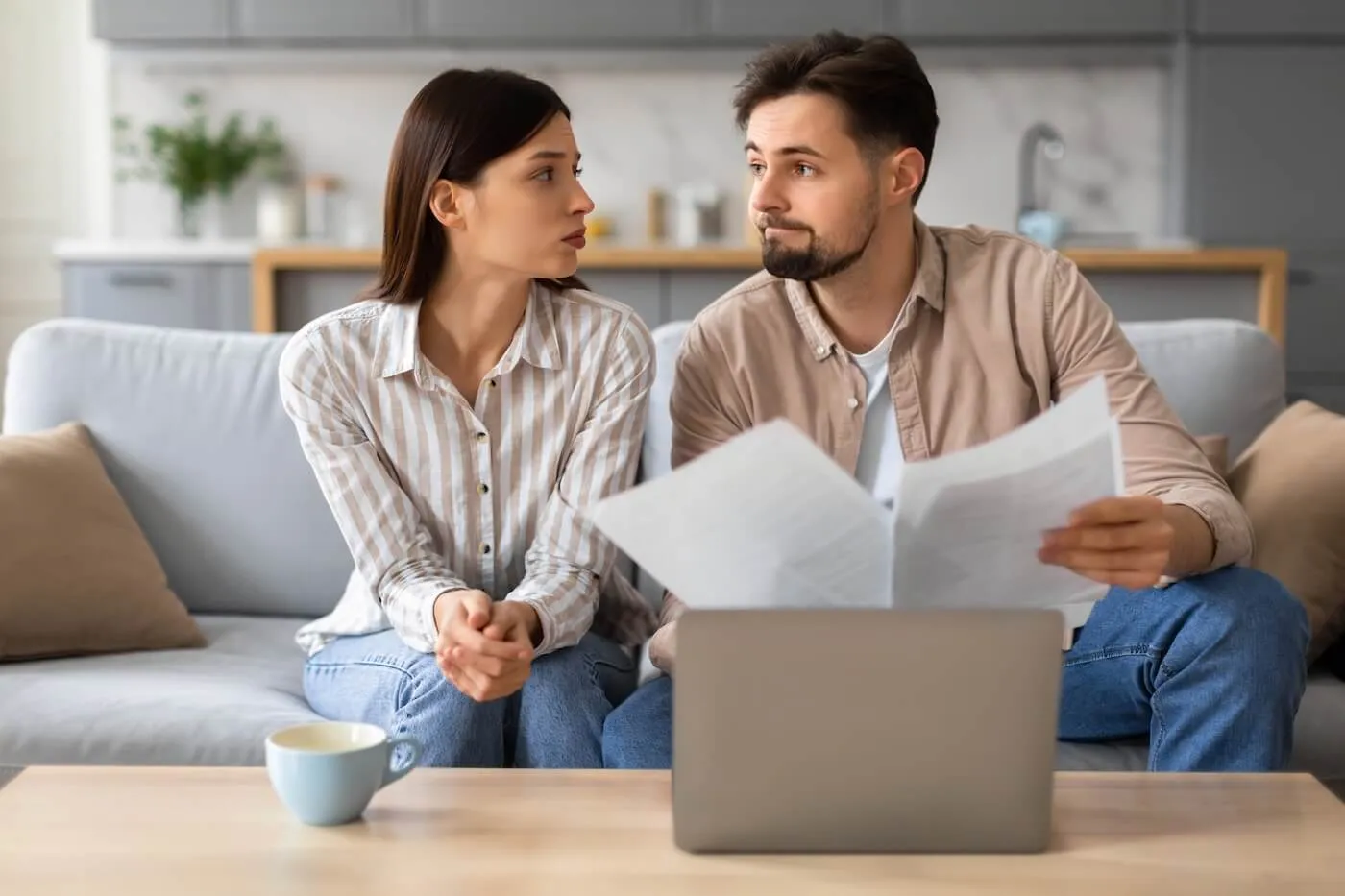 Concerned young couple examining paperwork with serious expression at home, with a laptop and a mug on coffee table.