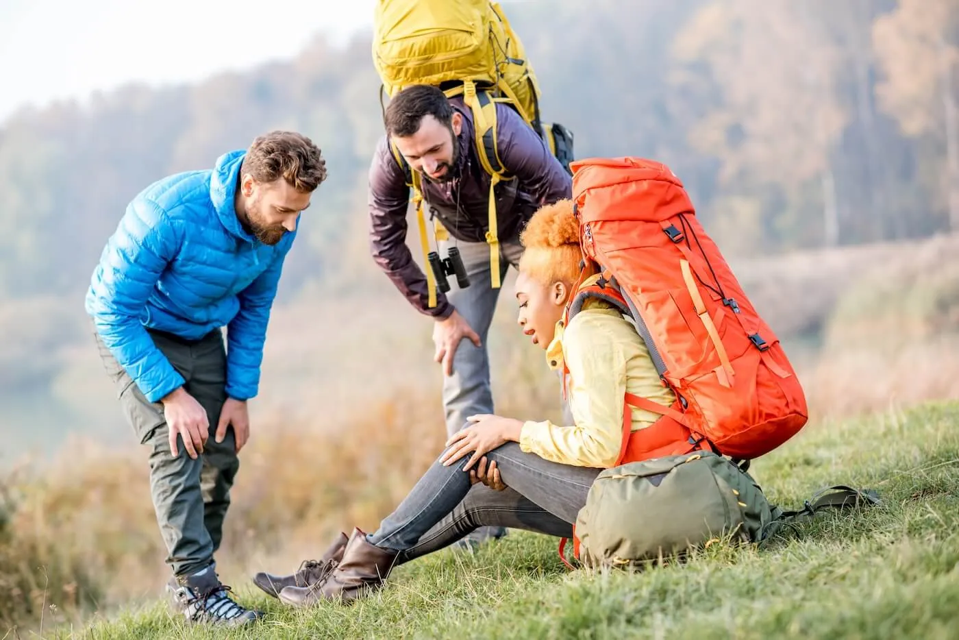 Young woman with a backpack sits on the ground with an injured knee while hiking in the mountains with two male friends
