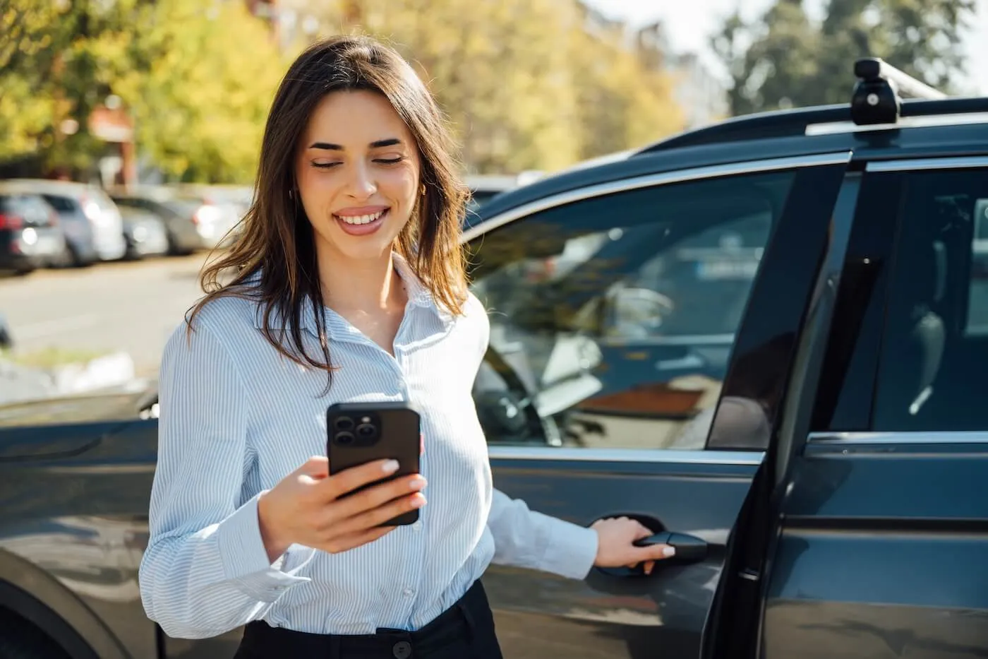 Smiling businesswoman standing in a parking lot, checking her smartphone while opening a car door