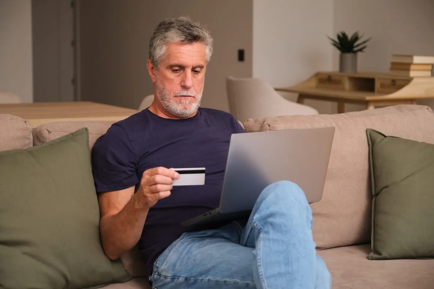 Focused mature man sitting on a sofa, holding a credit card while using his laptop