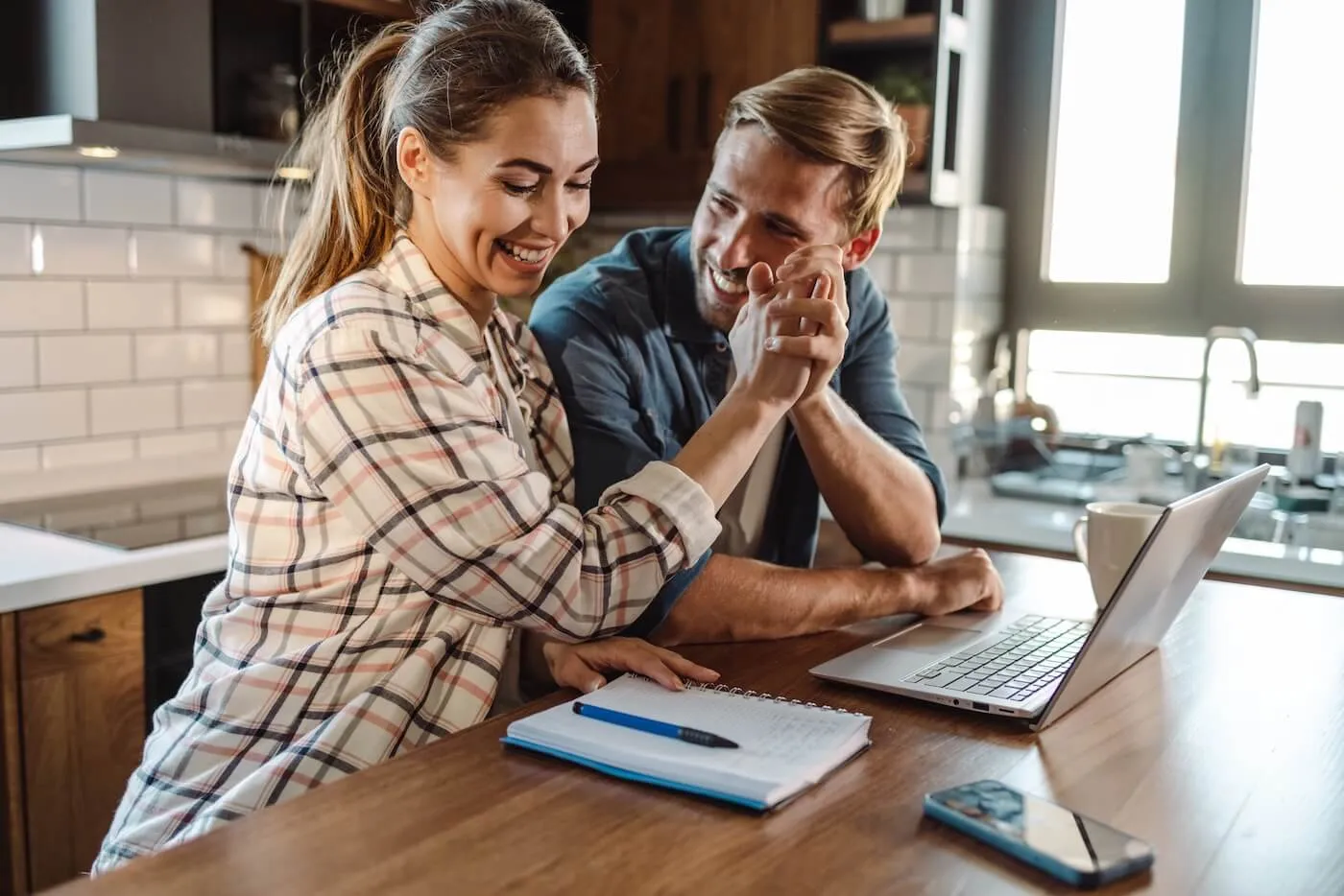 A cheerful couple sitting at a table at home, smiling and shaking hands while looking at a laptop, suggesting a successful submission of a mortgage application