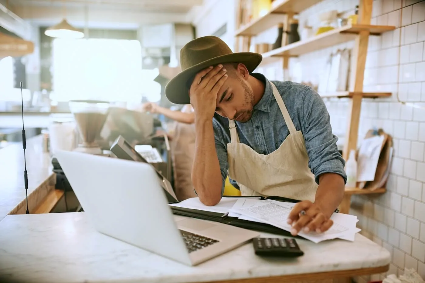 Small business owner seated at a café or restaurant counter, reviewing paperwork beside an open laptop and calculator. The workspace includes receipts, financial documents, and a point-of-sale area in the background.