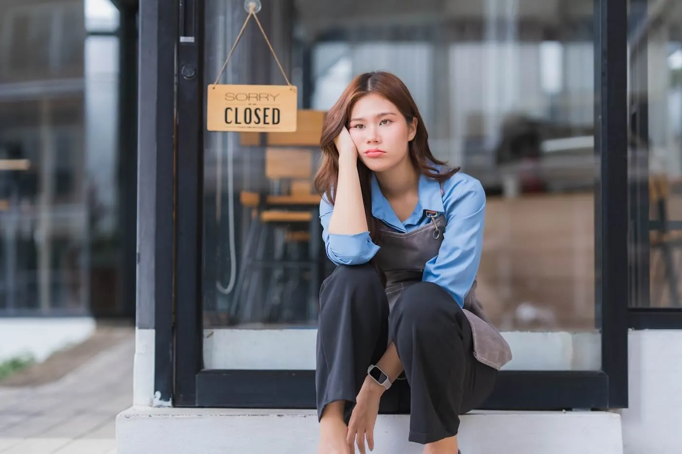 Small business owner sitting outside a storefront entrance beneath a sign that reads “Sorry Closed.” The upset woman is seated on a step in front of glass doors and windows, wearing a work apron and smartwatch, with the interior of the business visible in the background.