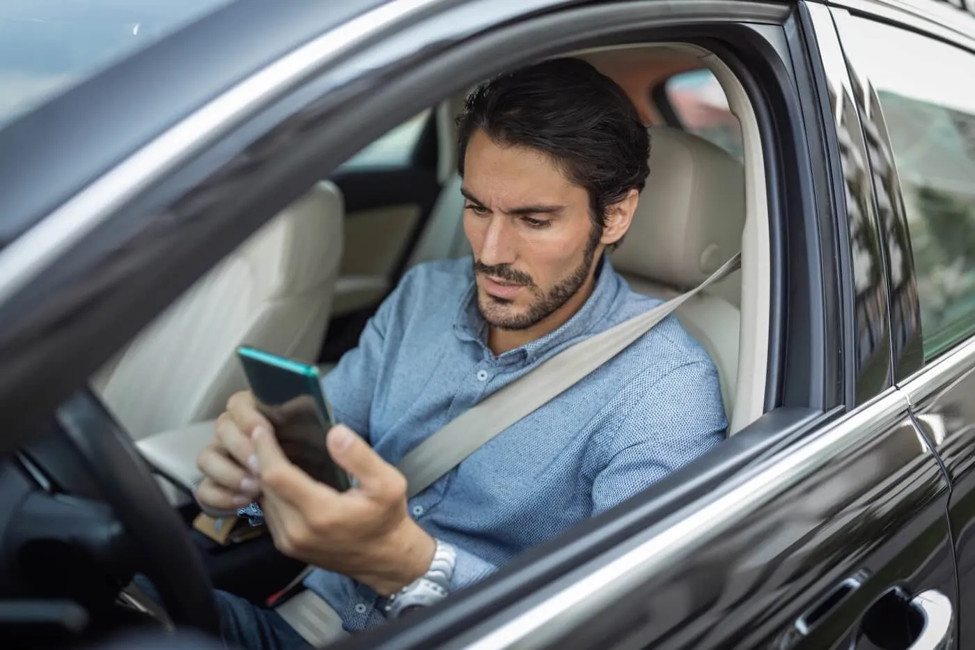 Focused man sits in the driver’s seat of a car wearing a seatbelt while looking at a smartphone.