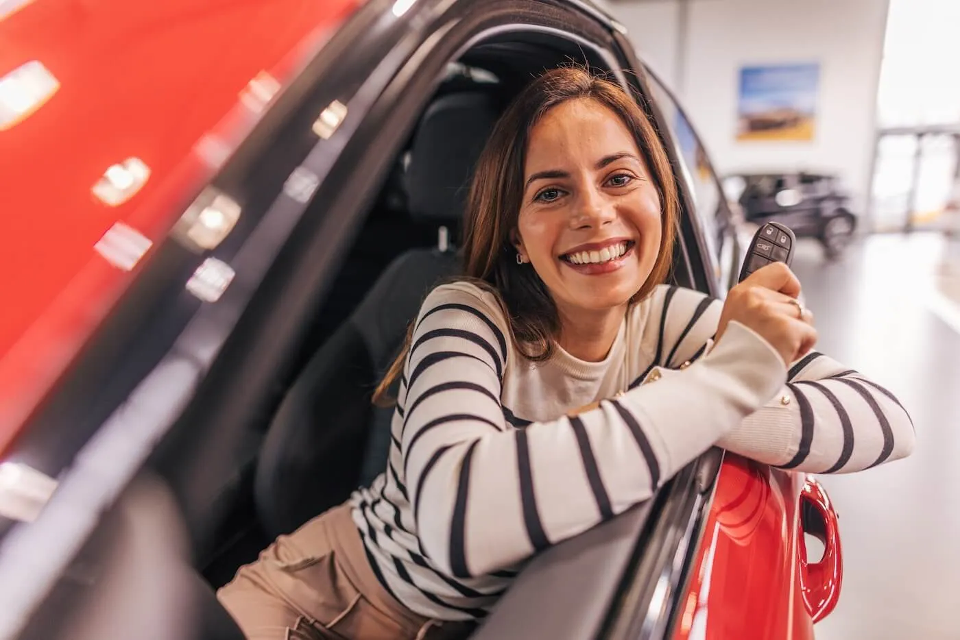 Photo of a happy young woman who is sitting in her new red car and holding car keys