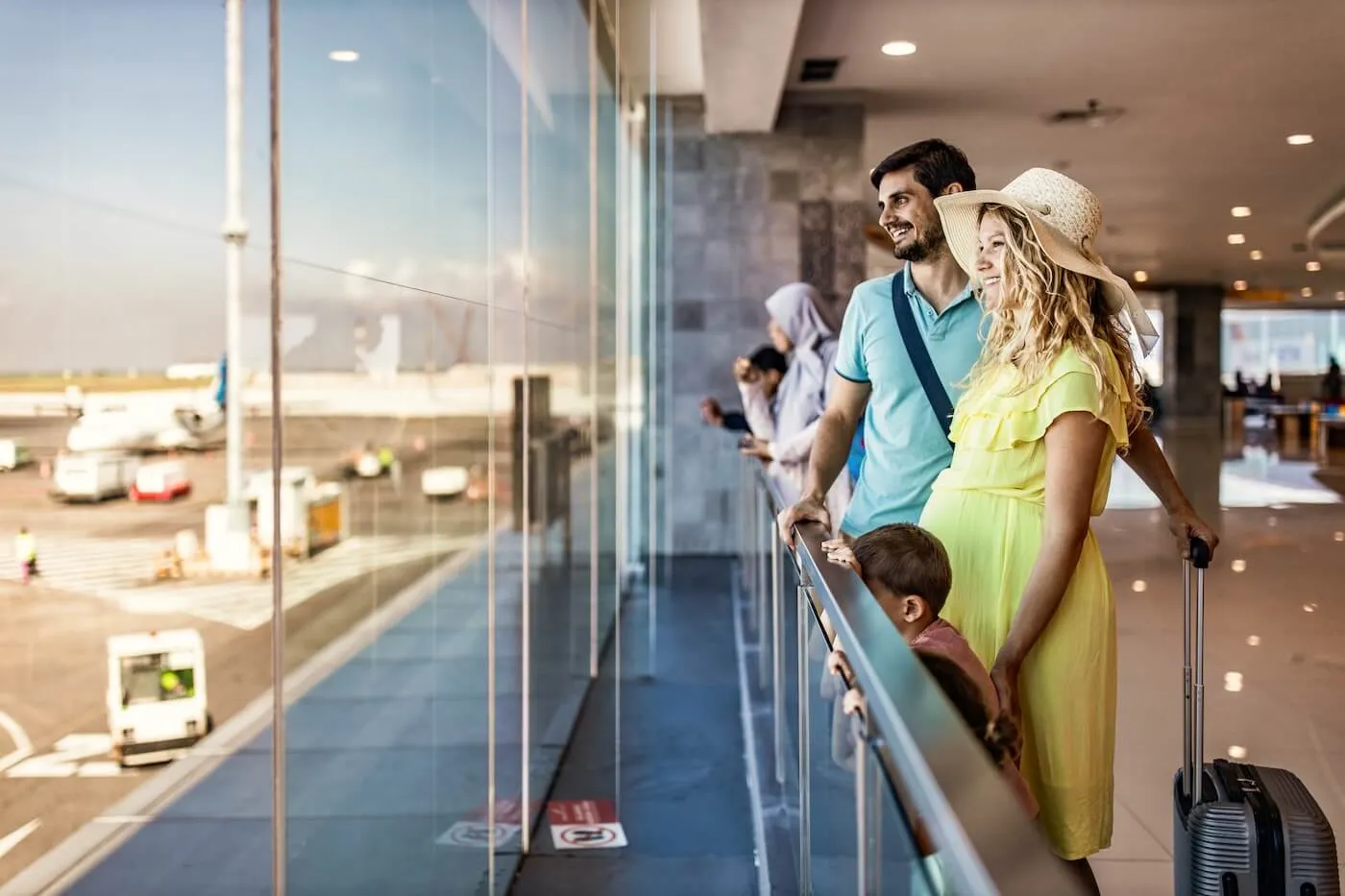 Family of three, including a pregnant woman in vacation attire, looking out an airport window at planes