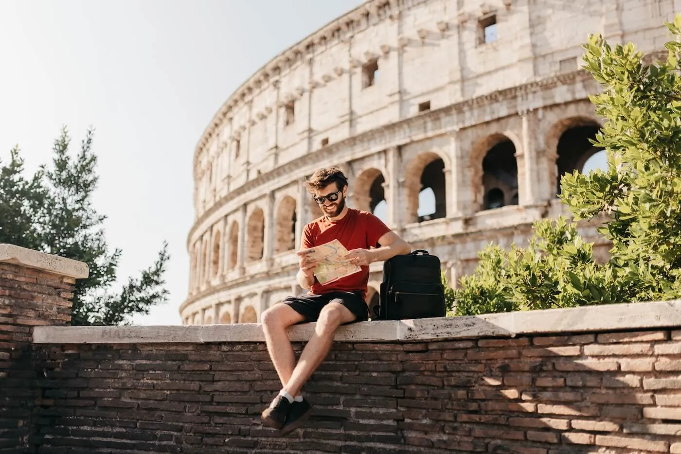 Smiling young man wearing sunglasses, sitting on a low wall near the Colosseum, exploring a map