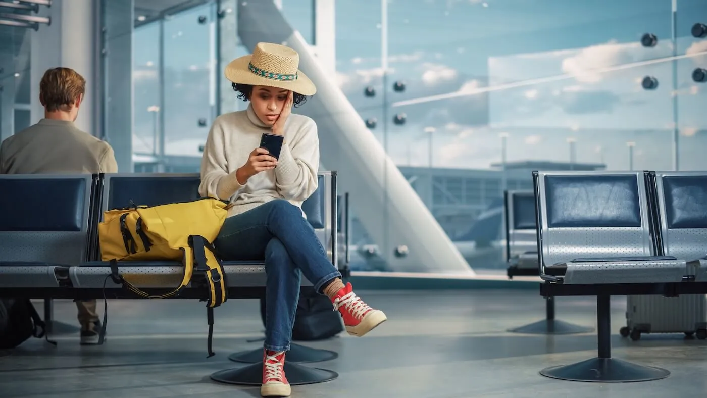 Frustrated young woman wearing a straw hat, sitting in an airport departure area with a yellow backpack beside her and checking her mobile phone