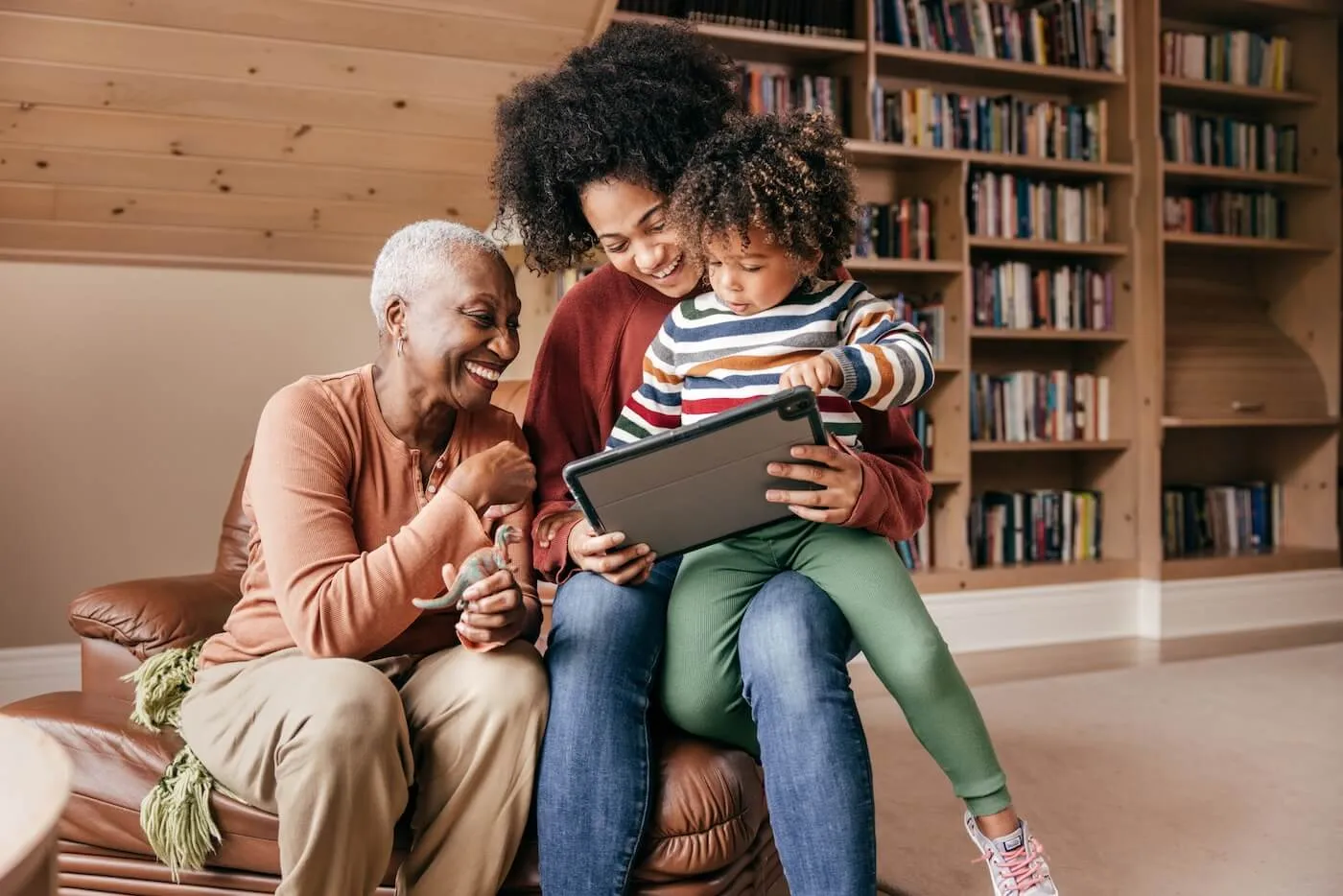 An elderly woman sits beside her daughter, who has a toddler on her lap; all three are gathered in a cozy home library, looking at a tablet together.
