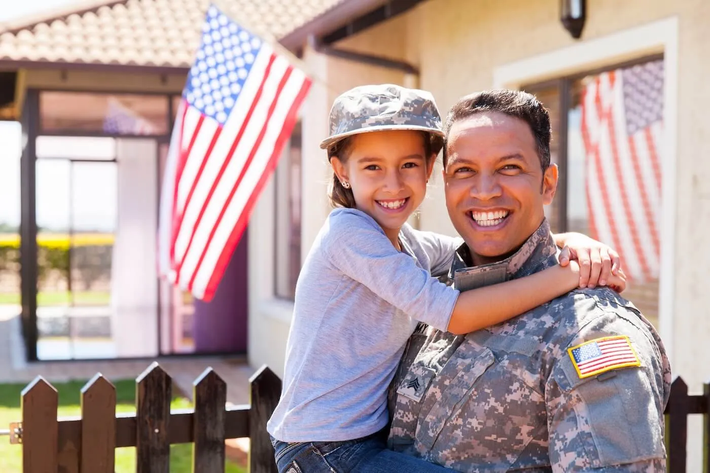 Smiling military man holding a girl who is wearing his cap; they are standing next to a house with the U.S. flag on it.