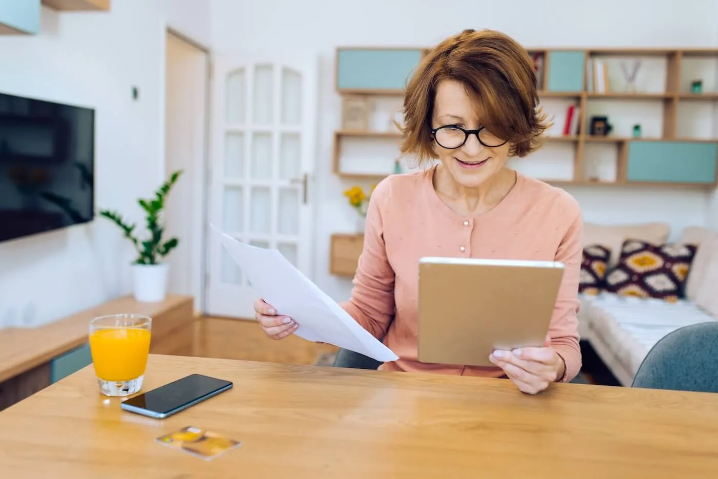 Focused mature woman comparing printouts with the data on her tablet at home