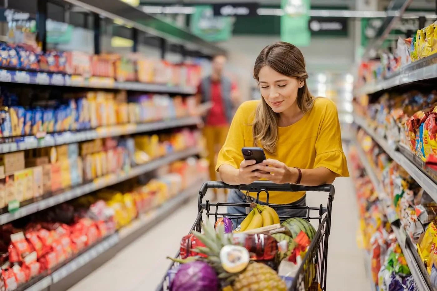 Smiling young woman checking her phone while pushing the full trolley in the grocery store