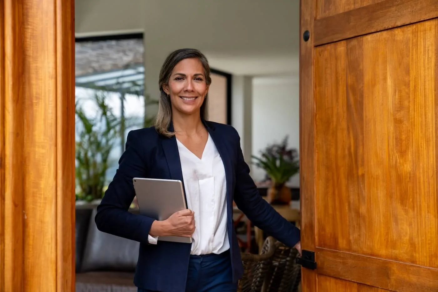 Female real estate agent with a tablet smiling while opening the front door to welcome visitors