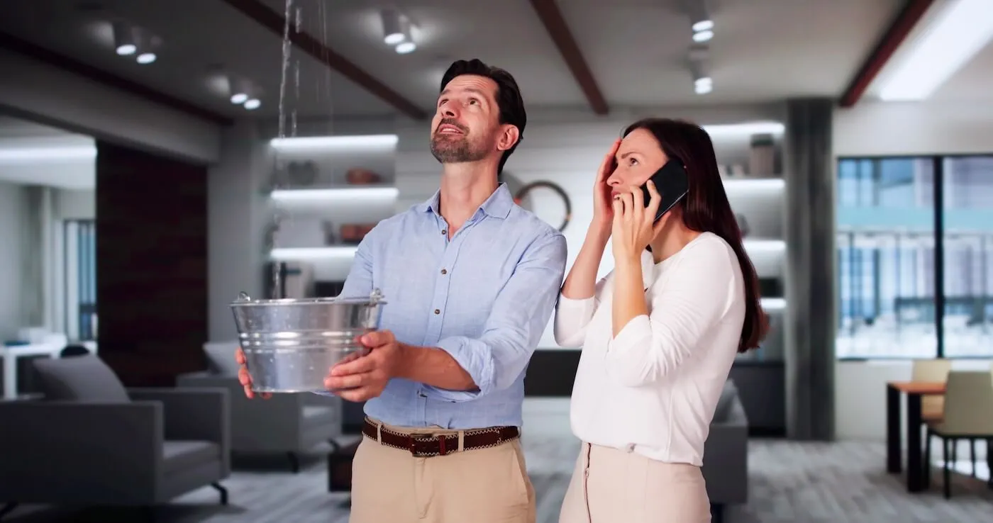 Concerned man holding a bucket under a ceiling leak while a woman nearby makes a phone call, likely seeking help for the emergency