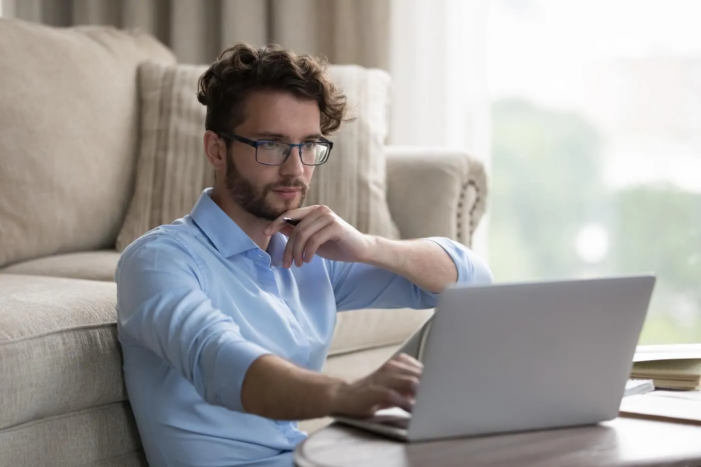 Focused young man using his laptop in a living room, sitting cozily next to a coffee table