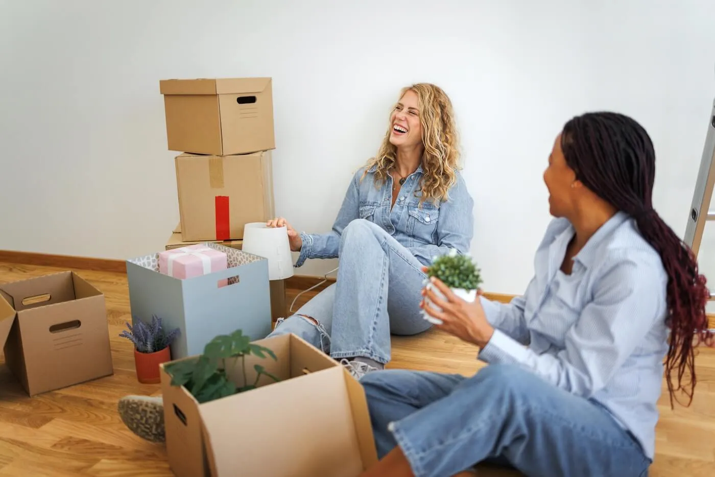 Two smiling women relaxing on the floor of their new apartment, surrounded by unpacked moving boxes, taking a break from settling in.