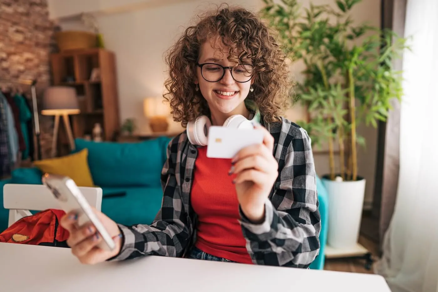 Smiling teenage girl with headphones around her neck holding a smartphone and using her credit card to shop online