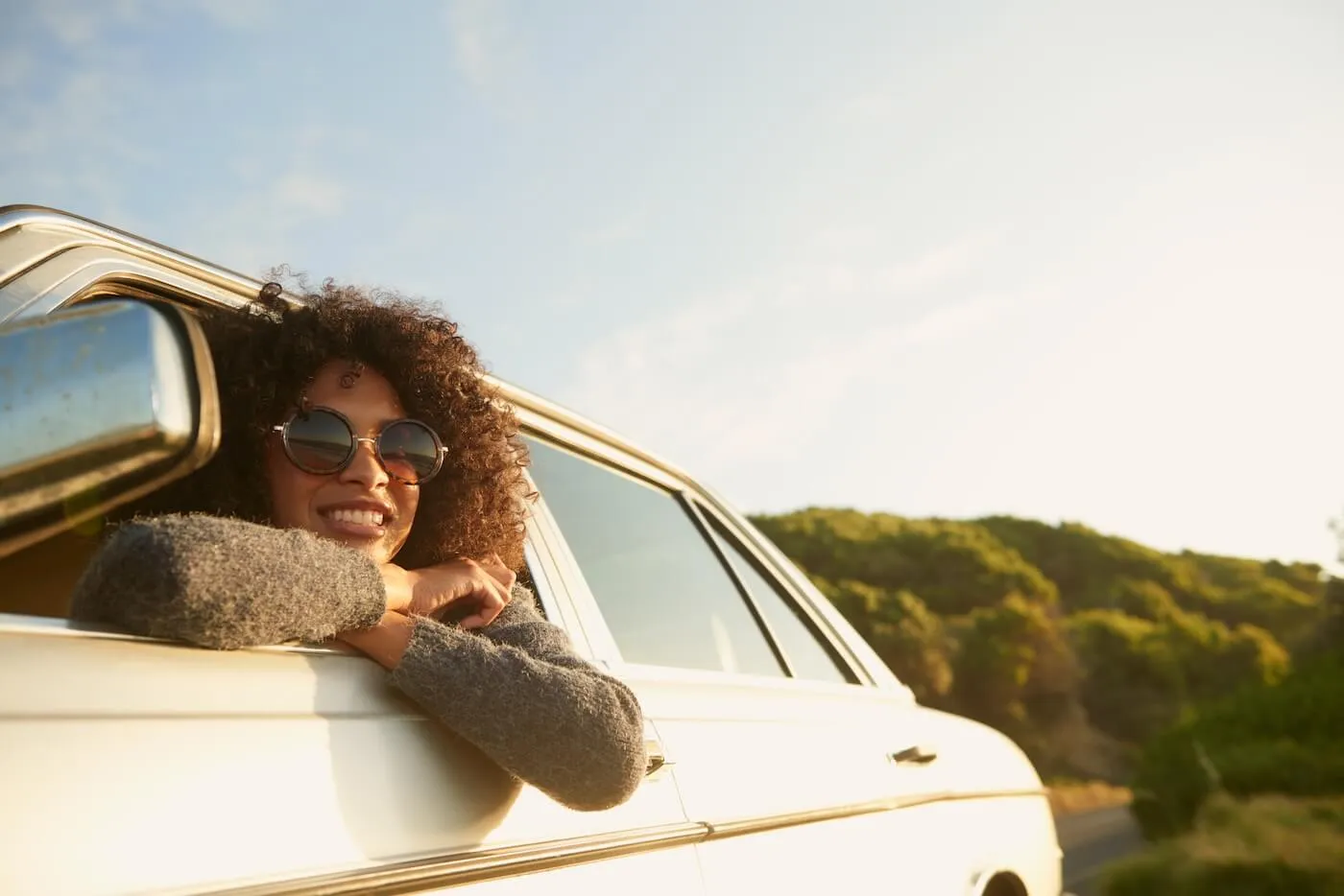 Smiling woman in sunglasses sticks out of the car window to enjoy the sunset