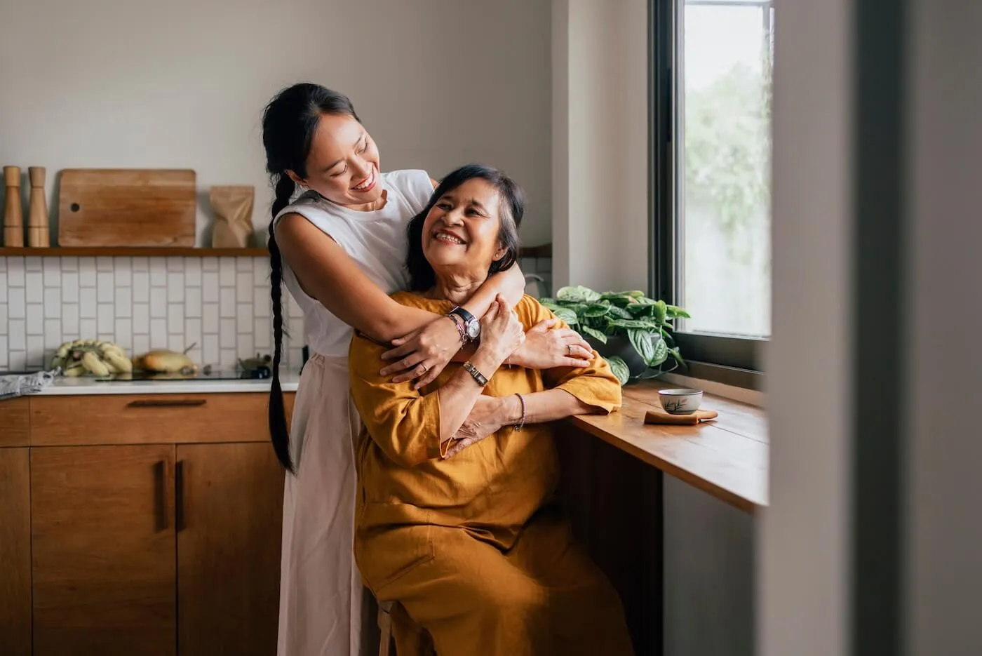 Smiling woman warmly embraces her elderly mother in a cozy kitchen, both appearing joyful and connected