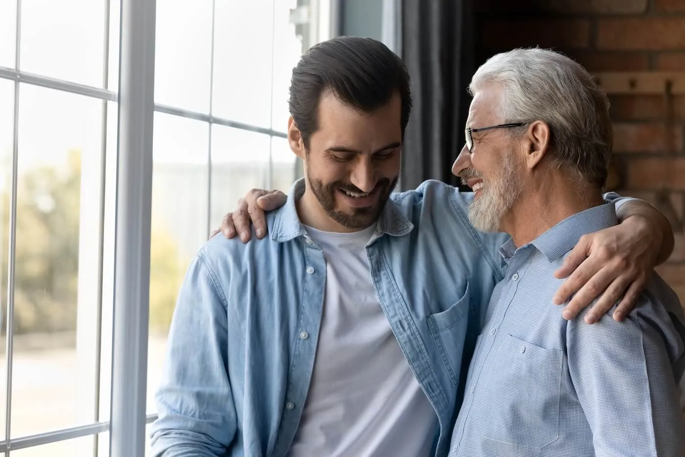 Smiling adult son hugging his elderly father as they stand together near a window