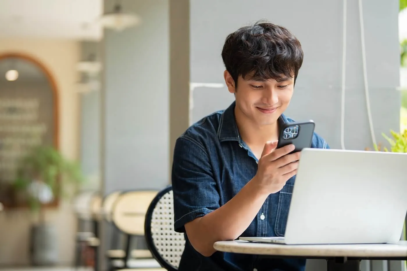 Smiling young man working at a café table, checking a smartphone while using a laptop