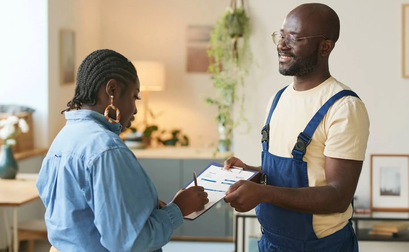 A smiling handyman wearing overalls hands an invoice to a female client for her signature.