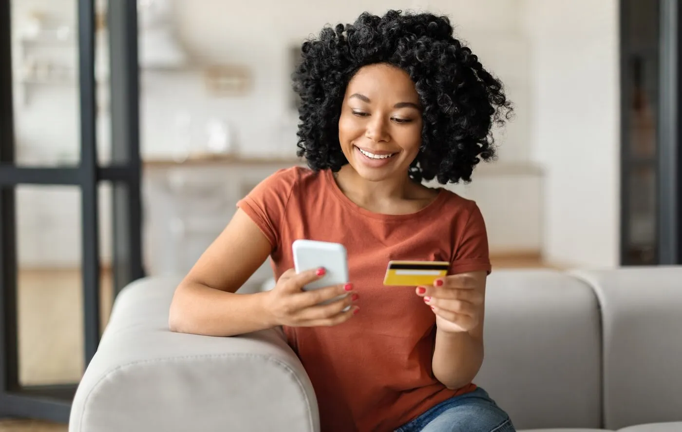 Smiling young woman sitting on a couch abd using her smartphone while holding a credit card