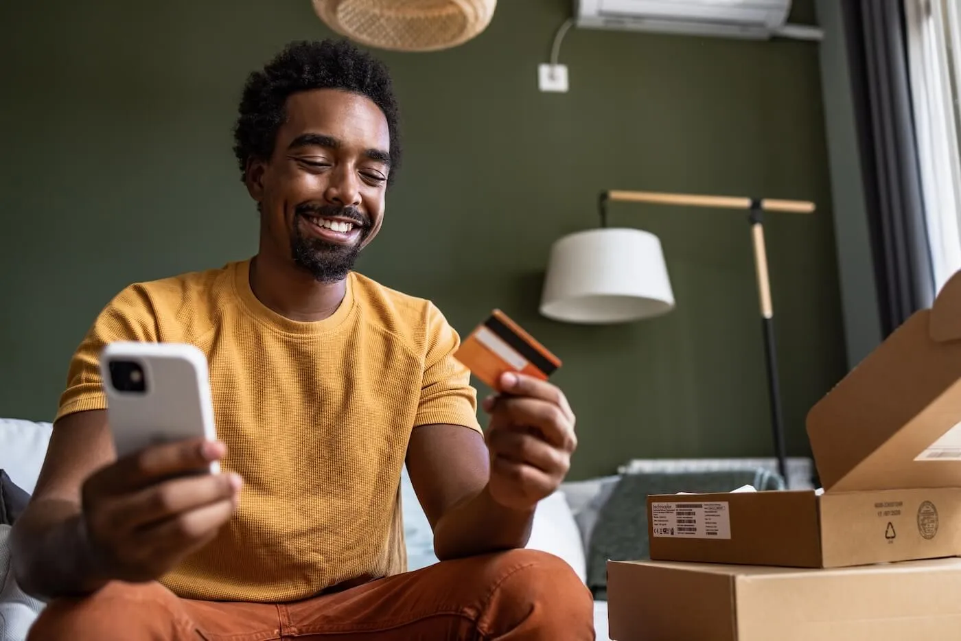 Cheerful man using his smartphone and holding a credit card, with shopping boxes beside him
