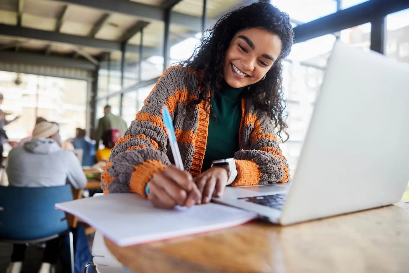 Smiling female student making notes in the classroom, with an open laptop beside her