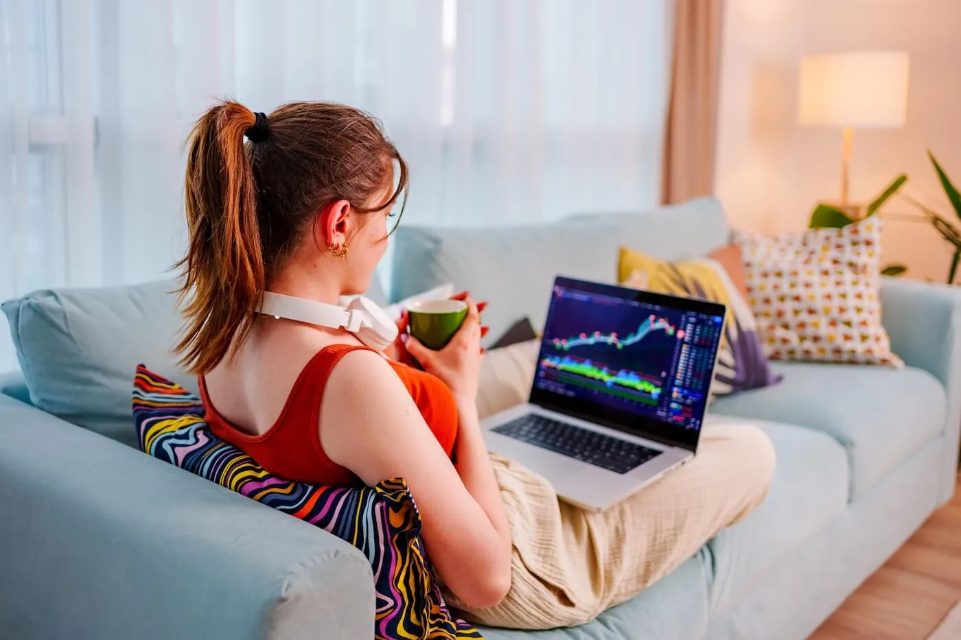 Young woman sitting on a couch with a laptop, checking stock market rates while enjoying a hot drink
