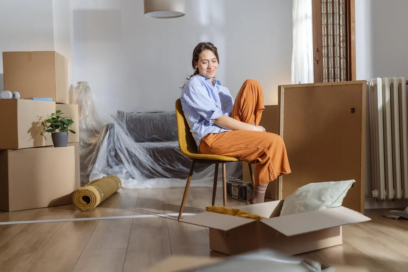 Young woman sitting comfortably on the chair in the middle of the room full of moving boxes and wrapped furniture