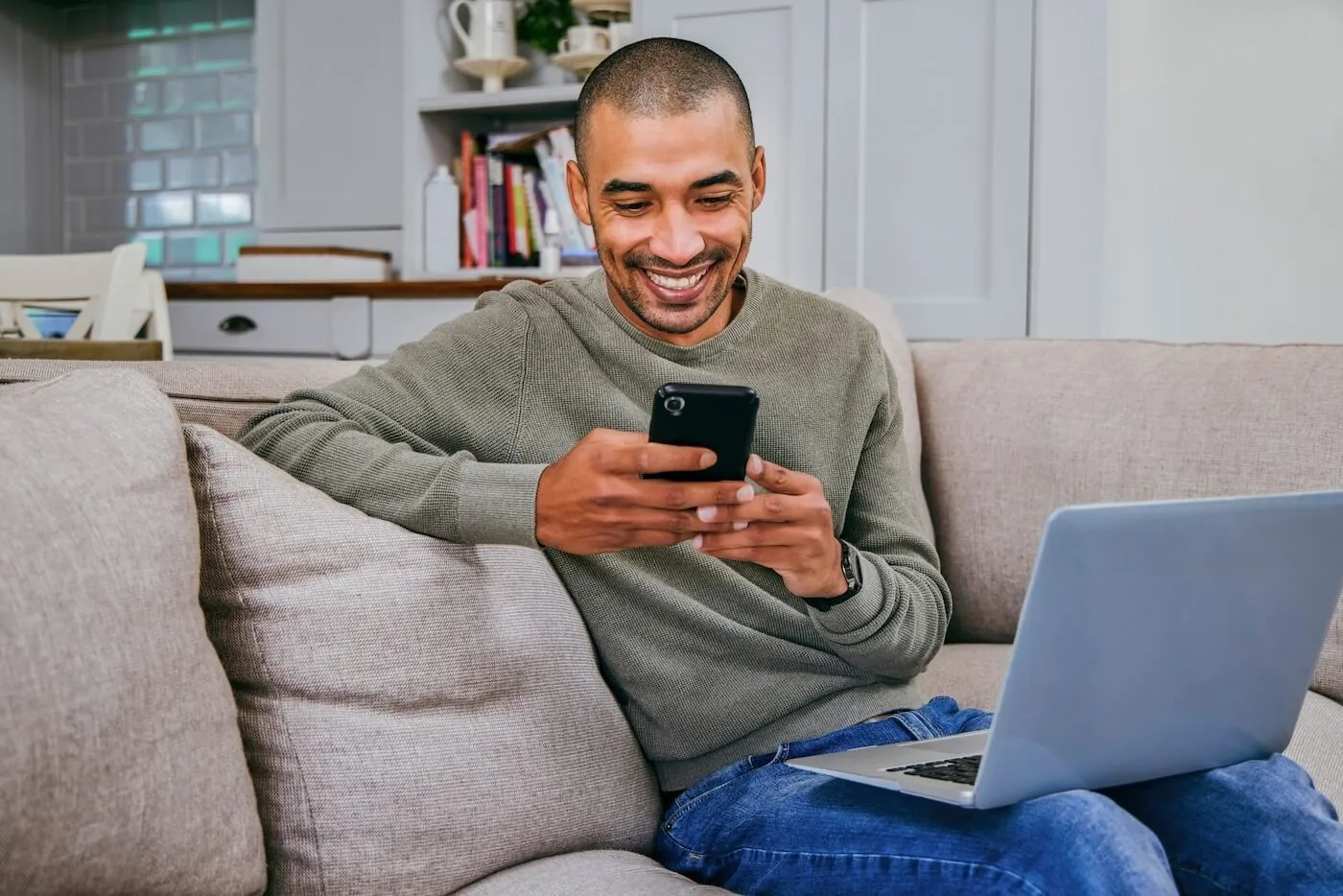 Smiling young man using his smartphone while sitting on a couch with an open laptop