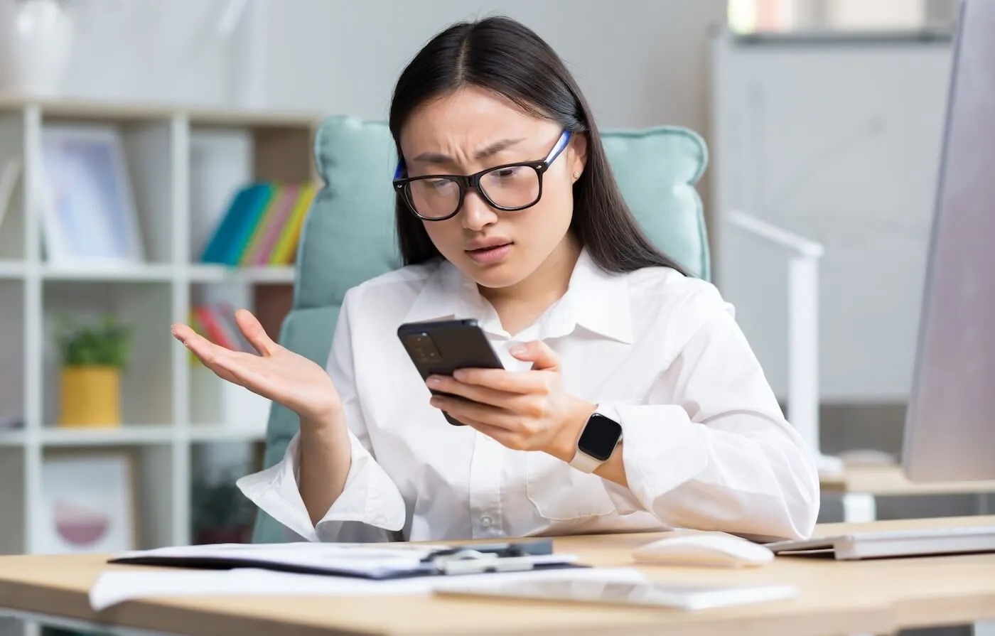 Concerned woman wearing glasses, looking at her smartphone while seated at a desk in an office setting