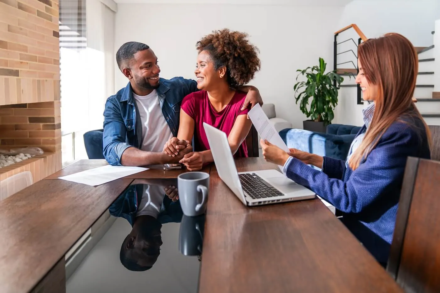 Happy couple sitting at the table with the female mortgage lender. Man and woman are holding hands and looking at each other while the smiling lender is reviewing the document, with a laptop next to her.