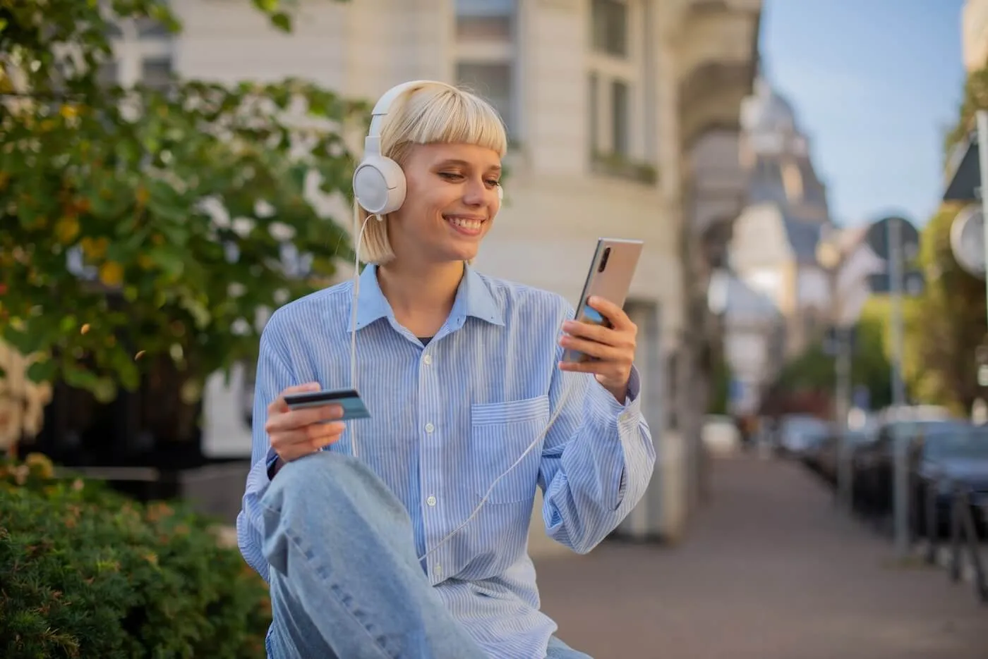 Smiling young woman with headphones using her smartphone and holding a credit card