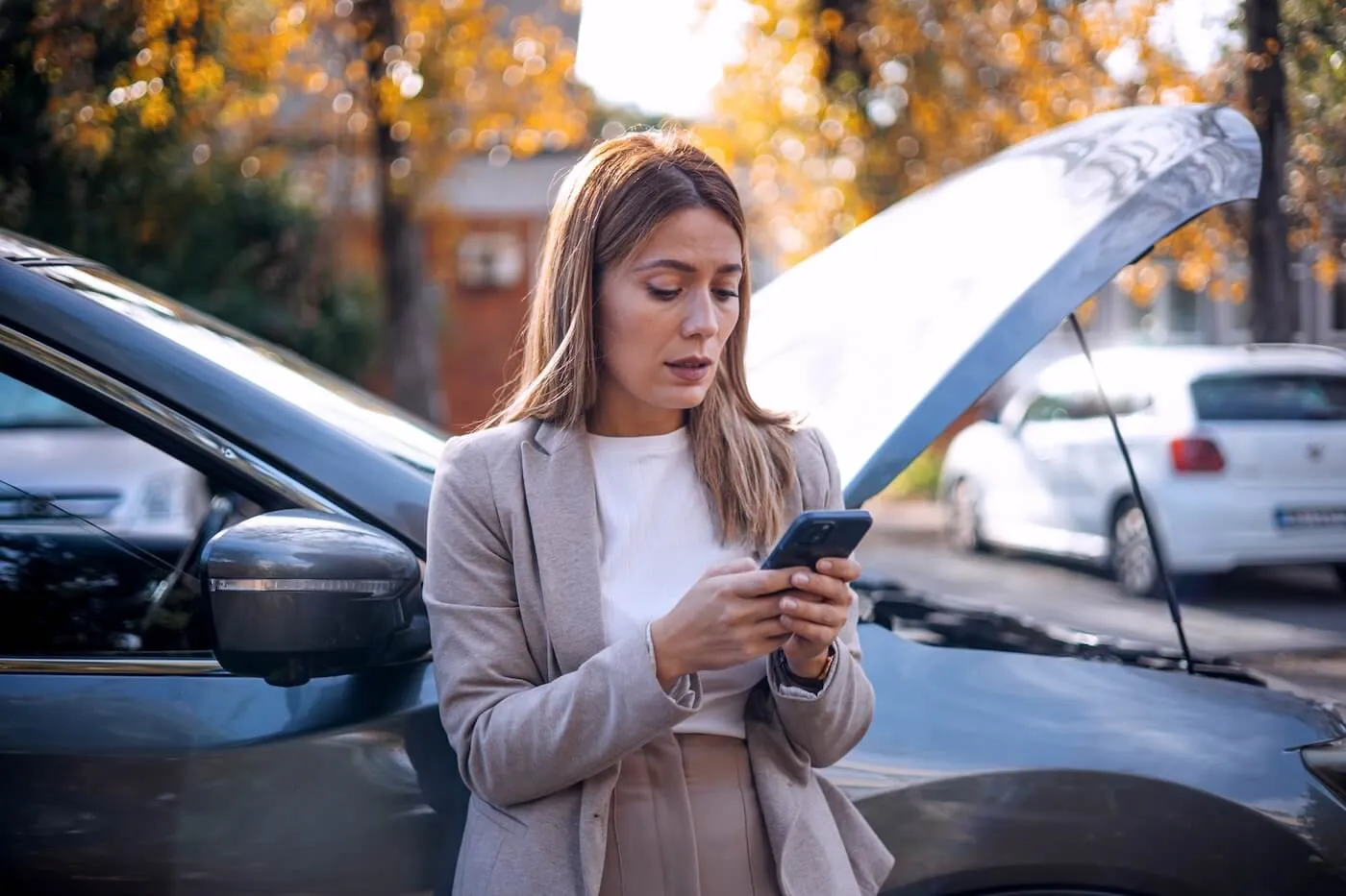 Concerned woman standing beside a broken car with the hood open, texting on her mobile phone