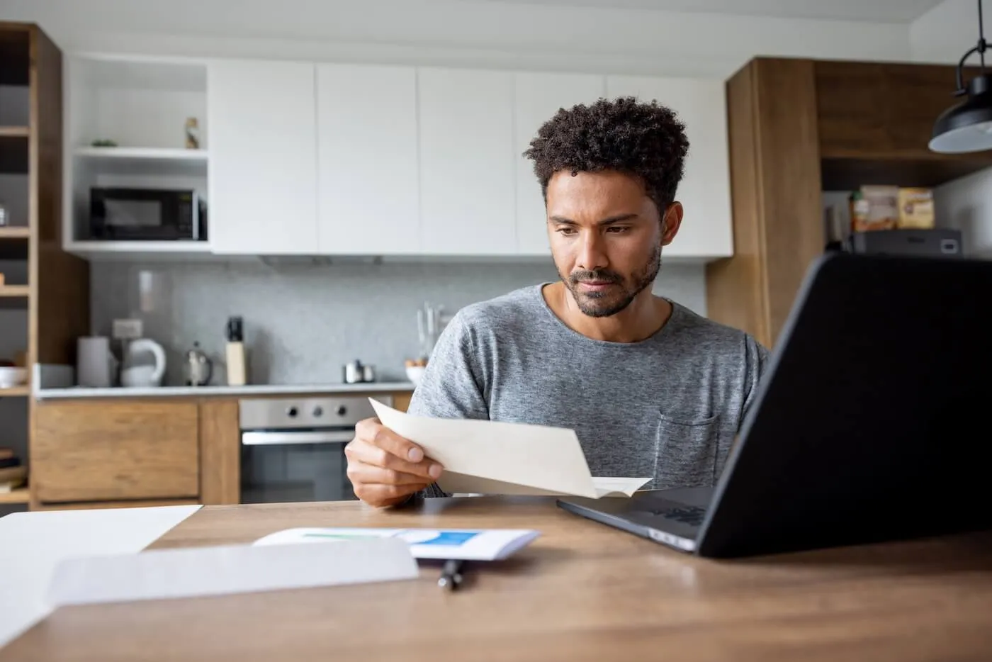 Focused man reading a letter while using his laptop in the kitchen