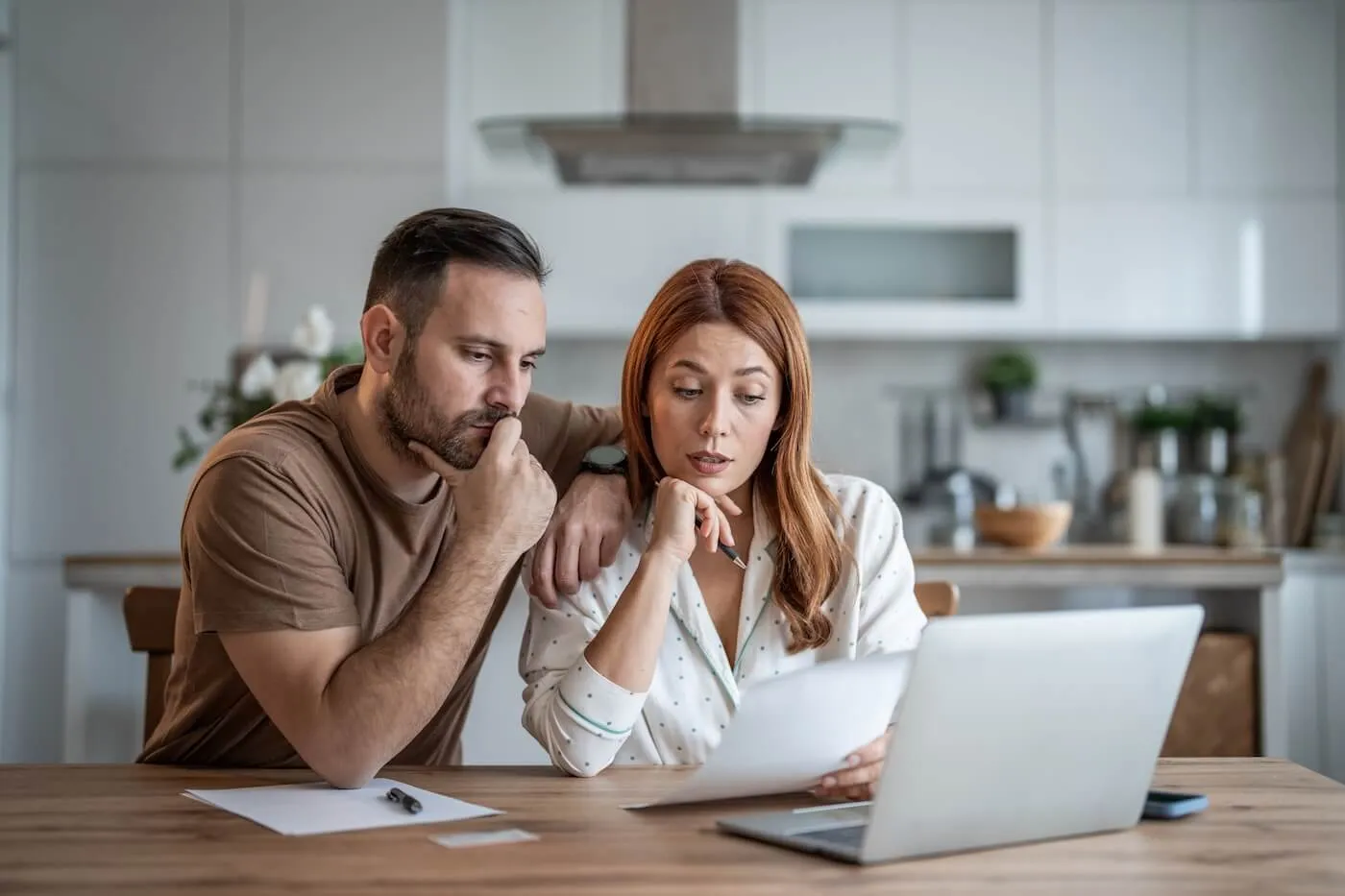 Concerned couple sitting together in the kitchen, reviewing a printed document with an open laptop beside them