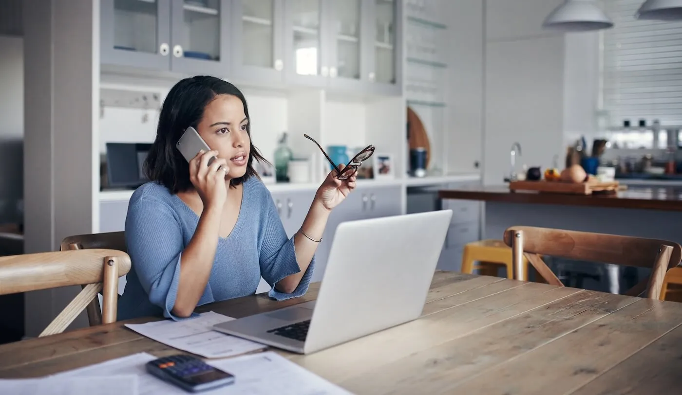 Focused young woman making a phone call while sitting at a dining table with a laptop, papers, a calculator, and glasses in hand