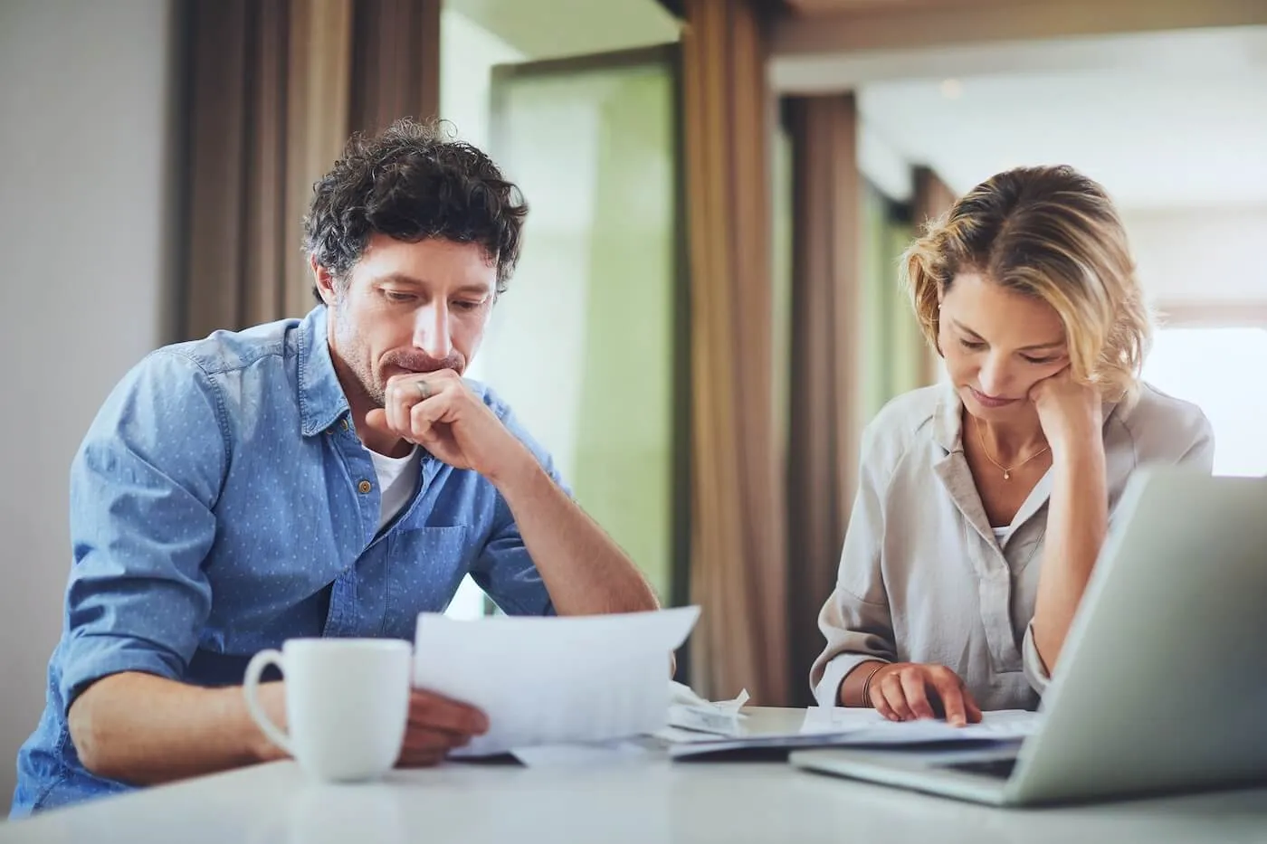 Focused mature couple reviewing financial documents in the kitchen with an open laptop and a mug beside them