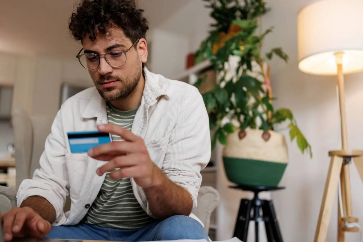 A person sits indoors holding a credit card while using a laptop, with papers on the table and a potted plant and floor lamp in the background.