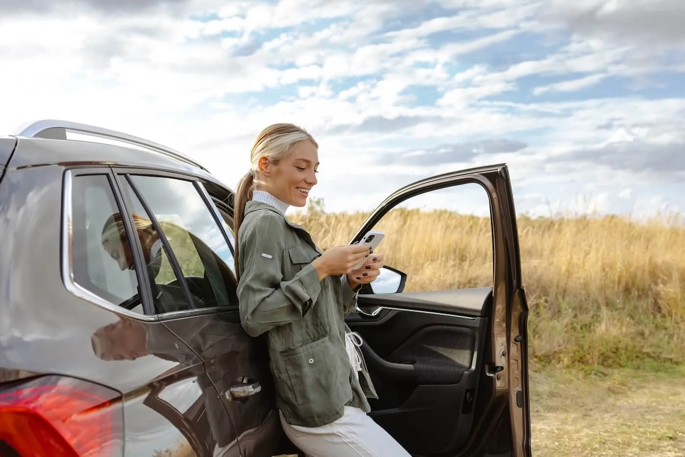 A smiling young woman standing in a countryside setting, leaning toward her parked car while checking her phone.