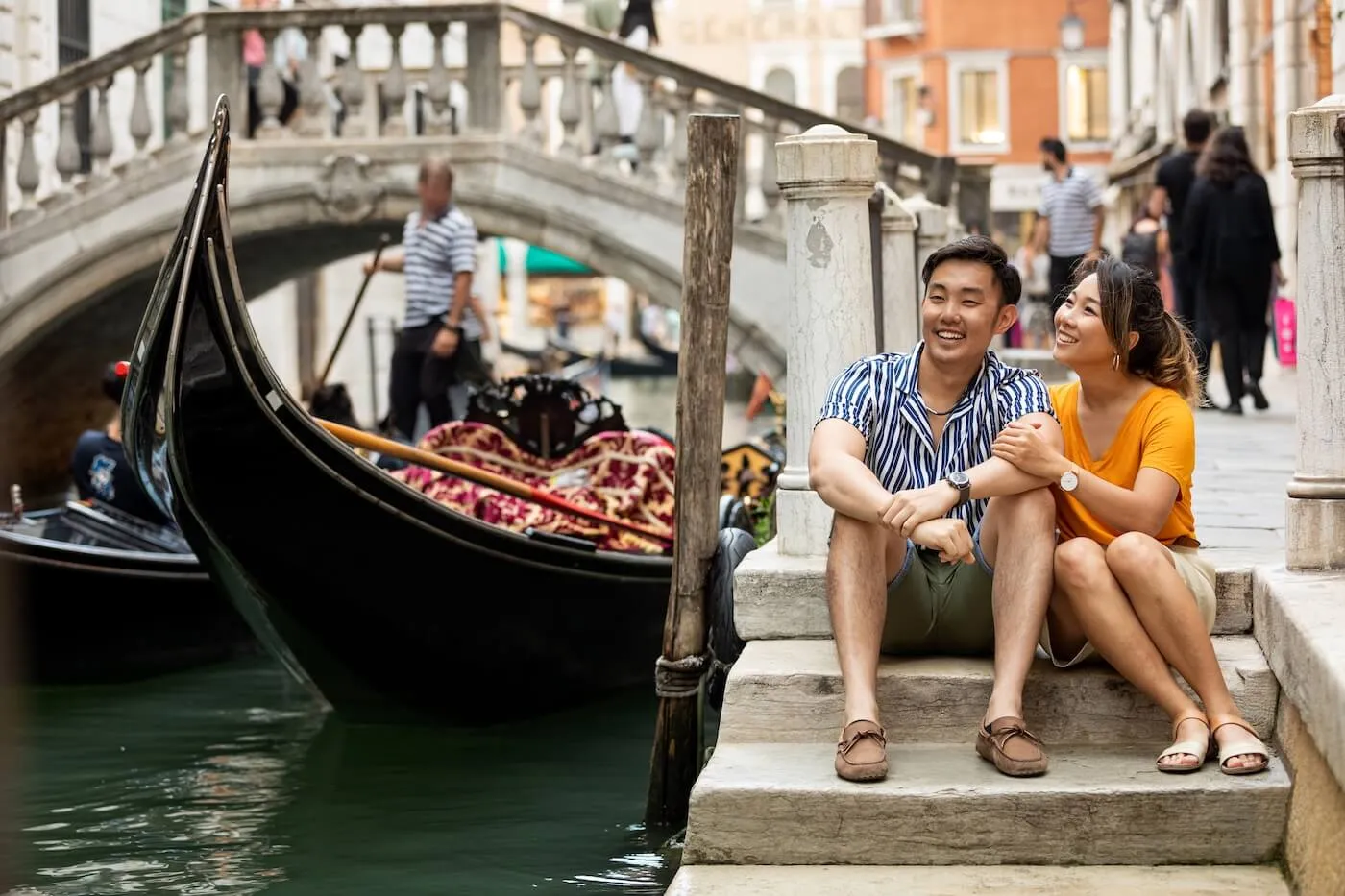 Happy young couple sitting on the stairs beside a gondola docked in a canal in Venice, enjoying the sunny day