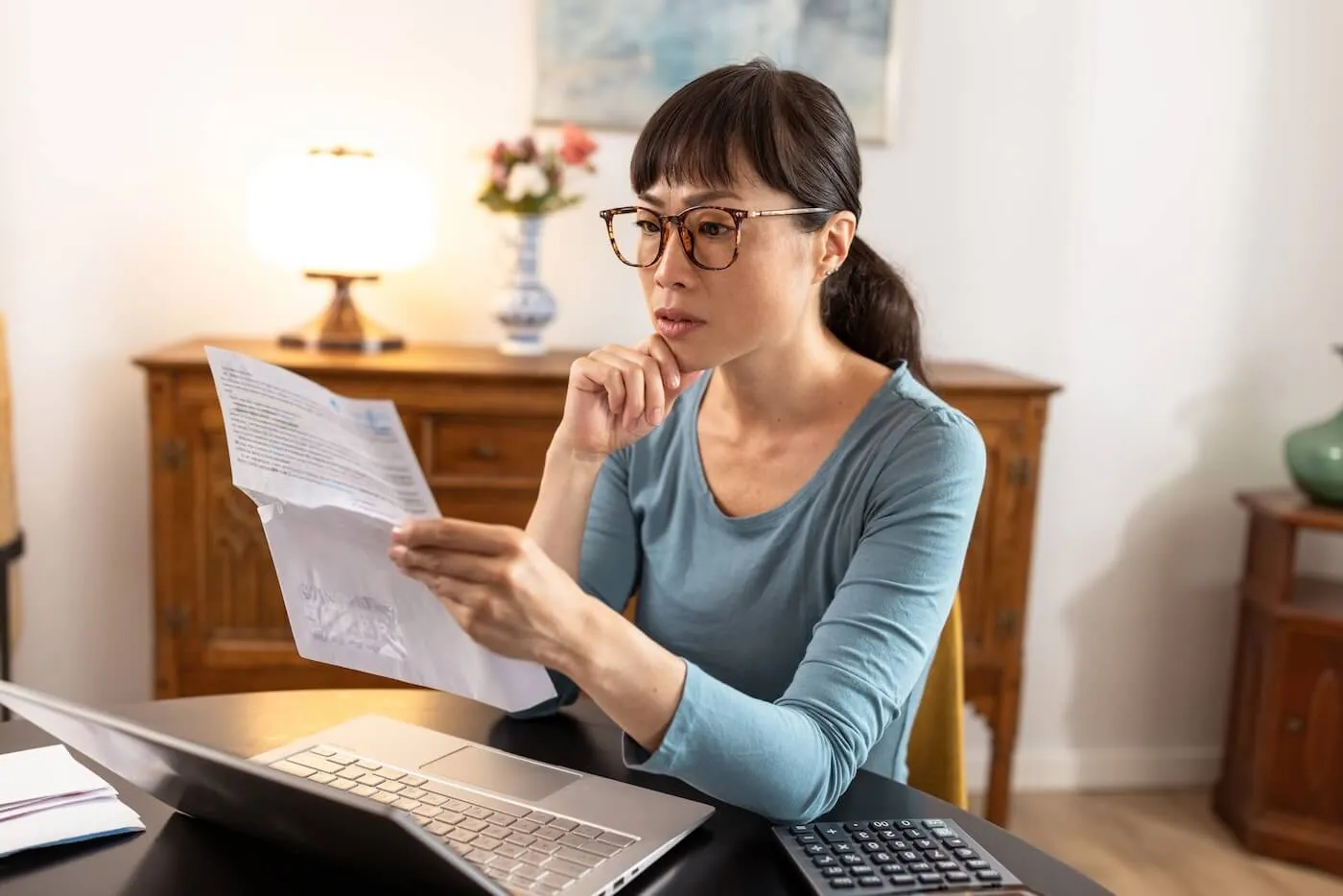 Concerned woman wearing glasses reviewing a bill at her home office desk