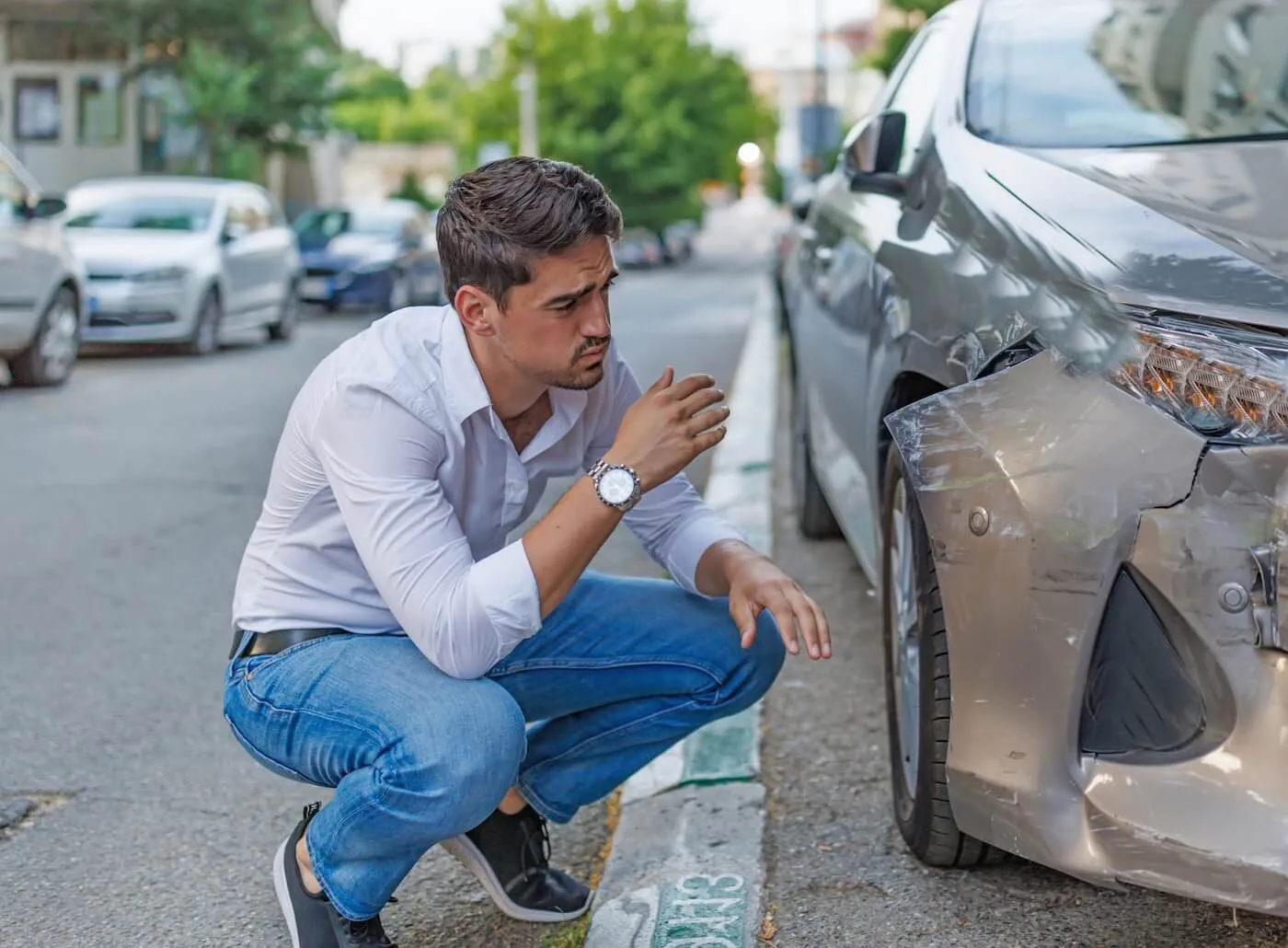 Frustrated young man crouching beside his damaged car, inspecting the extent of the damage on a city street