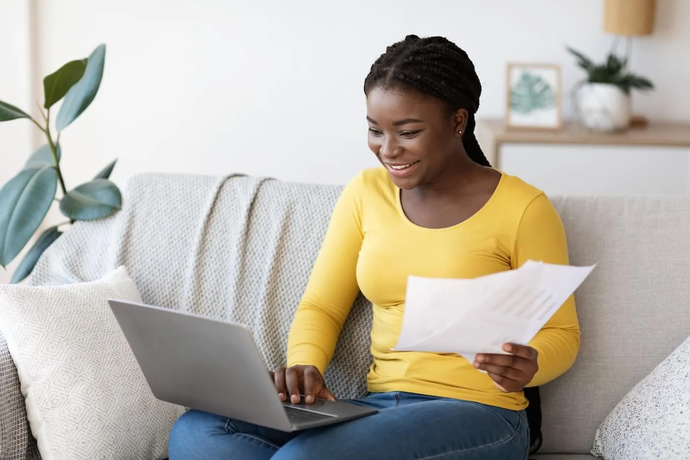 Smiling woman sitting on a sofa in a bright living room, holding financial documents while using a laptop. Houseplants, decorative furnishings, and natural light are visible in the background.