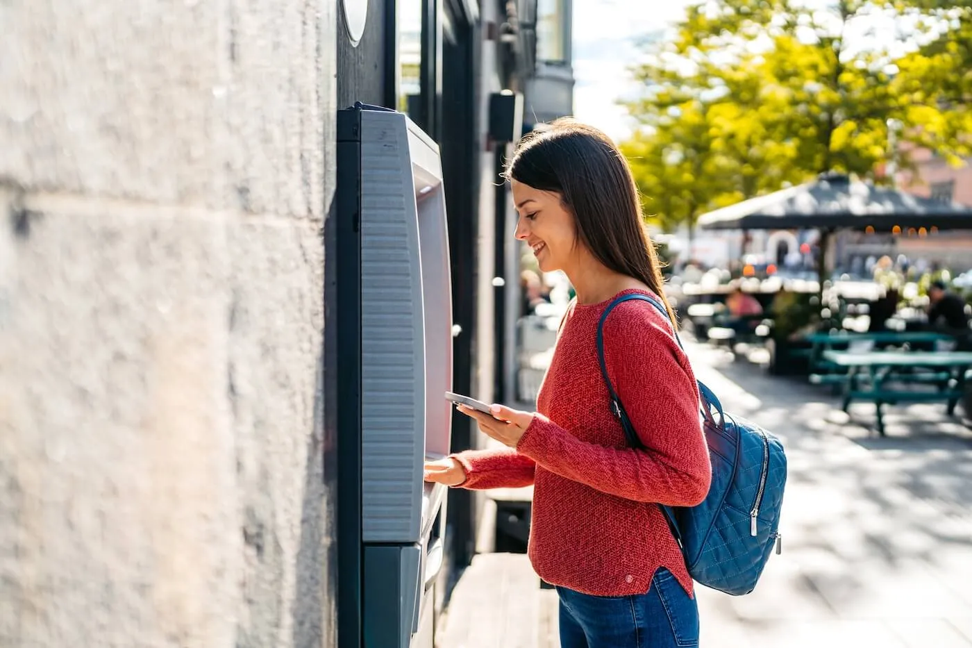 Smiling woman standing at an outdoor ATM, using the machine while holding a smartphone, with a backpack and a city streetscape visible in the background.