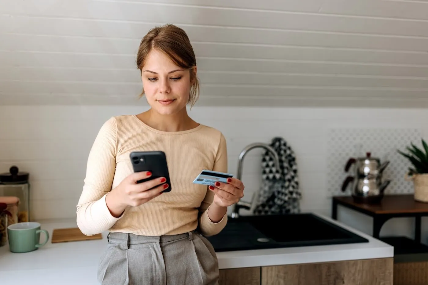 Focused young woman standing in a modern kitchen holding a smartphone in one hand and a credit card in the other. The image illustrates online shopping, mobile banking, or digital payments, with a sink, countertop, mug, and kitchen appliances visible in the background.
