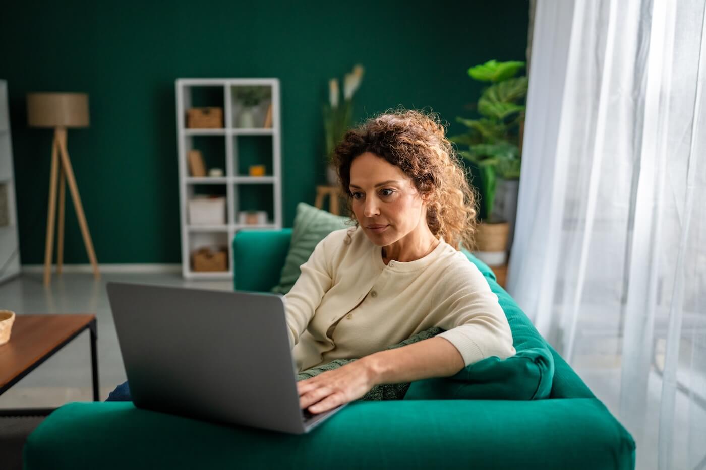 Focused woman sitting on a green sofa using a laptop in a modern living room, with plants, shelving, and large windows providing natural light in the background.