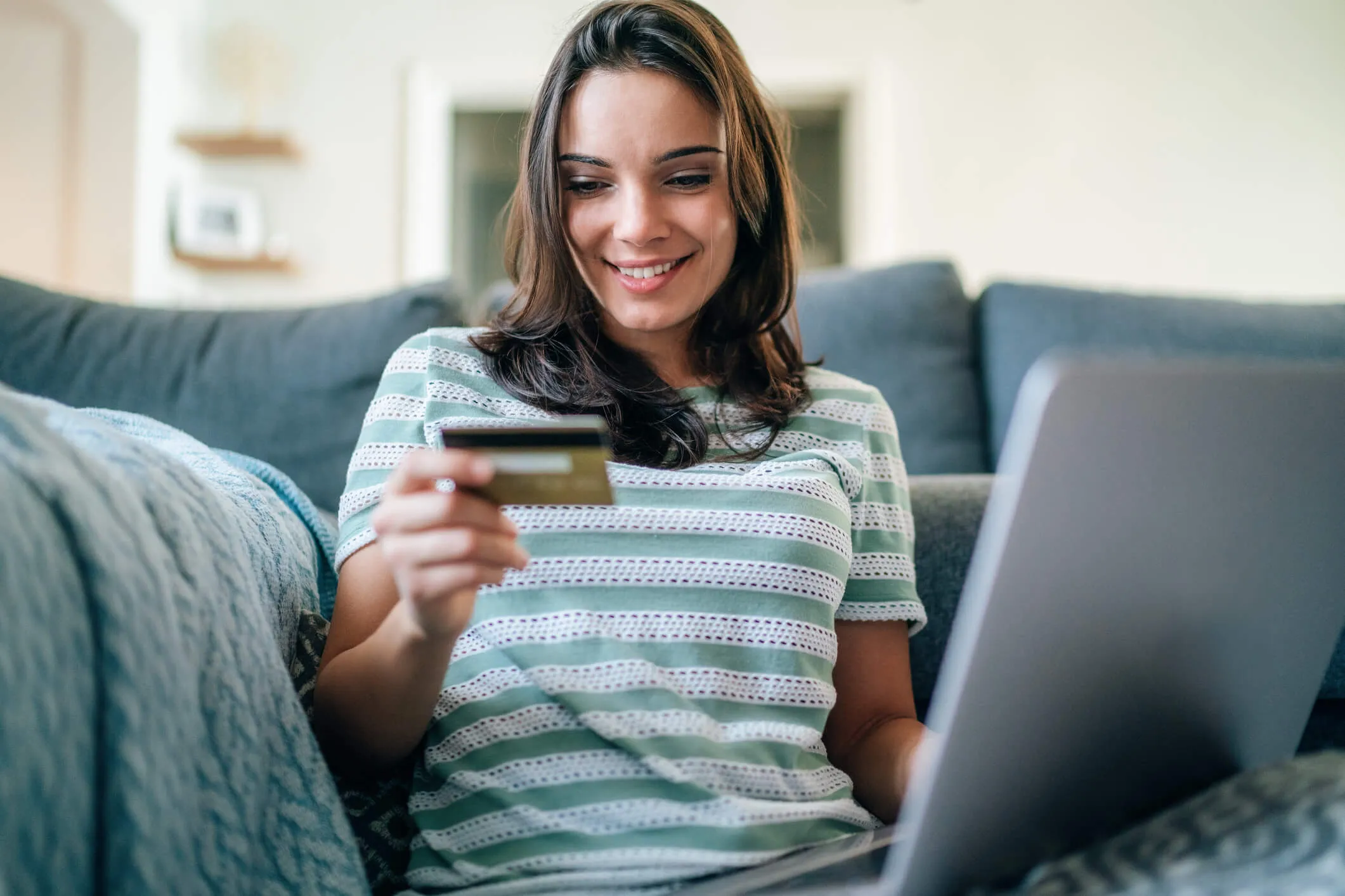 Smiling young woman sitting on a sofa at home using a laptop while holding a credit card, making an online purchase in a comfortable living room setting.